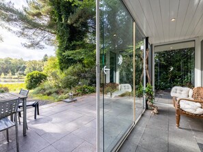 Een moderne buitenpatio met een glazen wand, een houten bank en een tafel met stoelen.