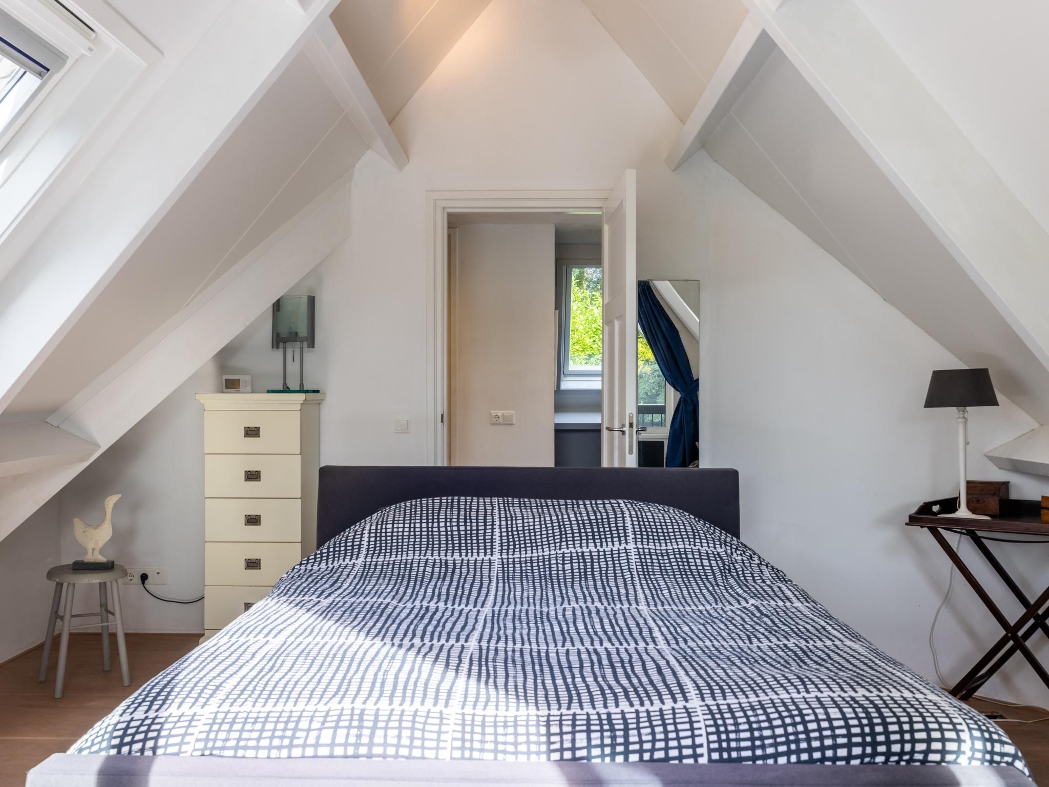 A bedroom with a sloped ceiling, a bed with a patterned bedspread, a dresser, a lamp, and a window.
