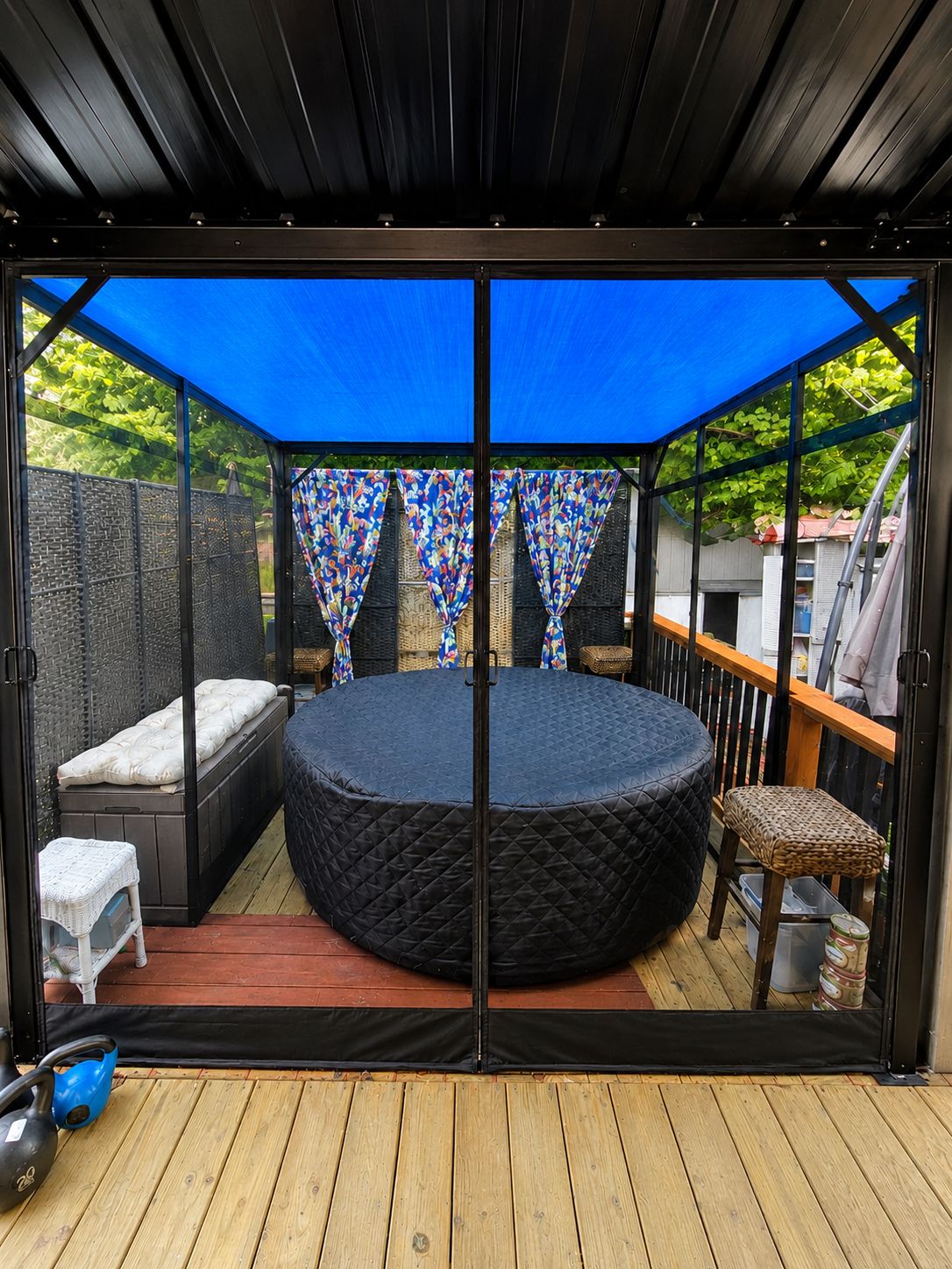 Un espacio de estar al aire libre circular con una mesa redonda y bancos, rodeado por una terraza de madera y un toldo azul.
