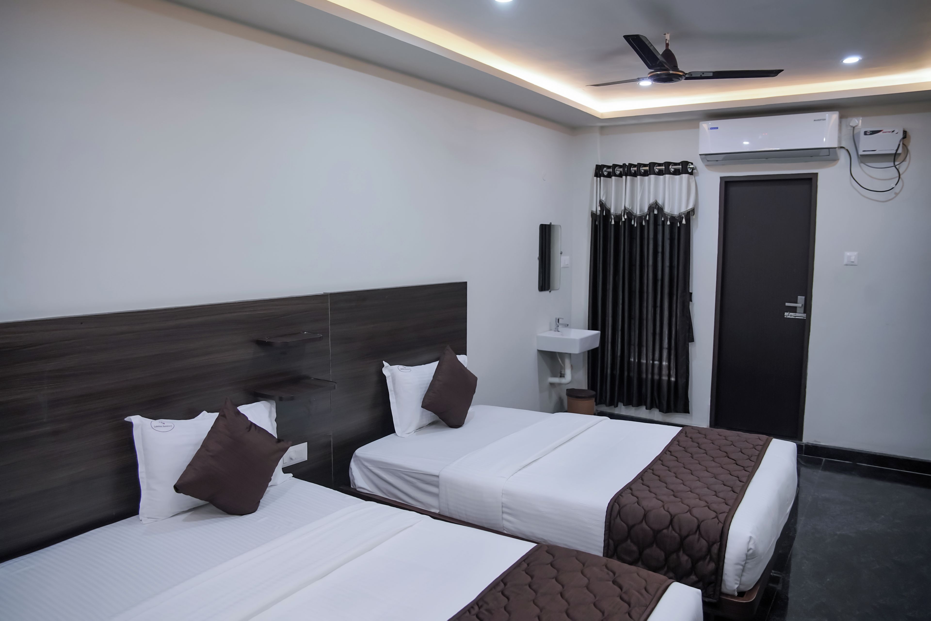 Habitación de hotel con dos camas, ventilador de techo, unidad de aire acondicionado, baño con lavamanos y una puerta.