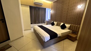 A modern hotel room with a large bed, a bedside table, an air conditioning unit, and a view of the outside through a window with curtains.