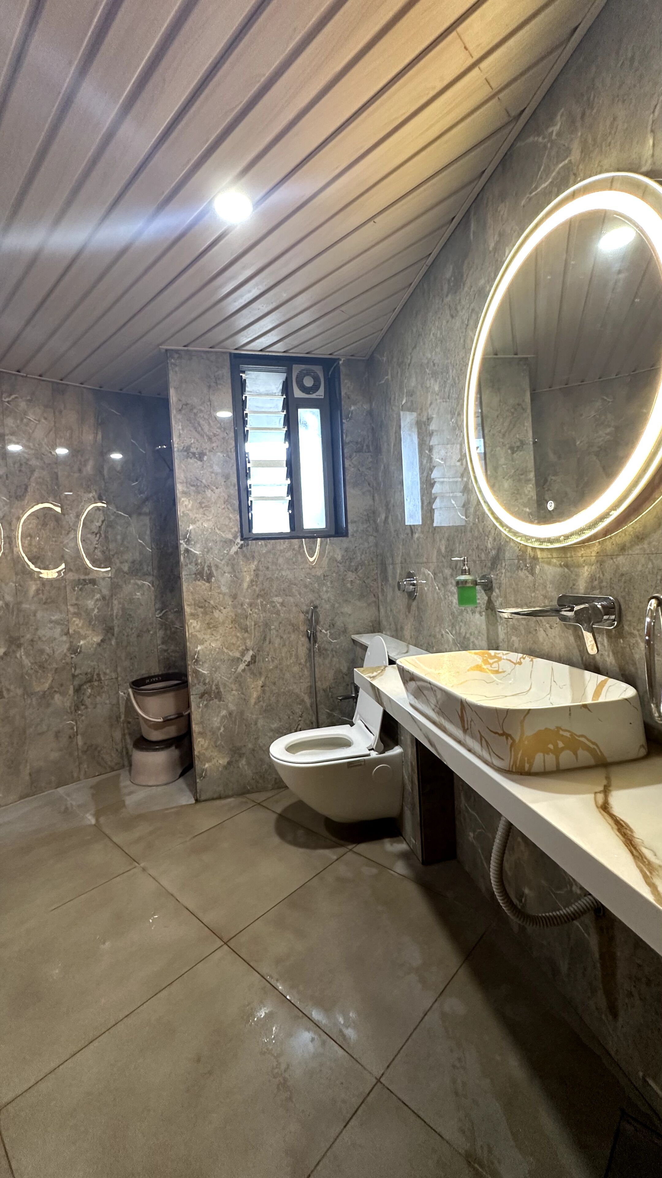 A bathroom with a marble wall, a toilet, a sink with a mirror, and a window.