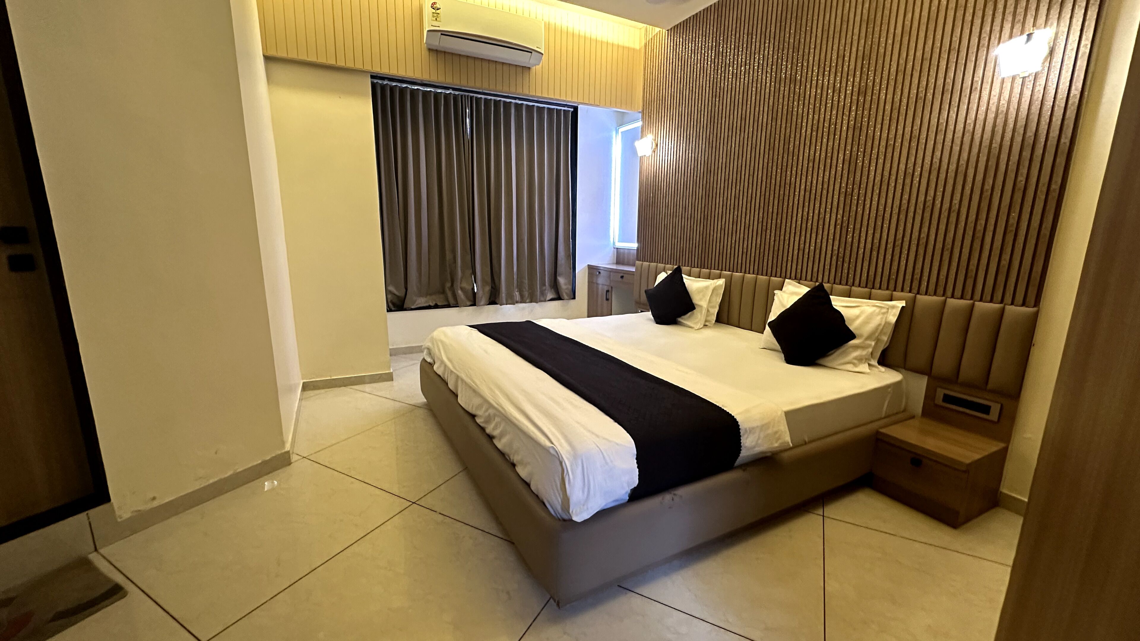 Habitación de hotel moderna con una cama grande, una mesita de noche y un ventanal con cortinas.