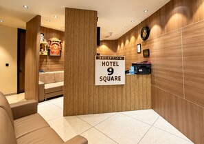 Zona de recepción con mostrador, un cartel que indica 'RECEPTION HOTEL 9 SQUARE', un sofá y una pared con paneles de madera vertical.
