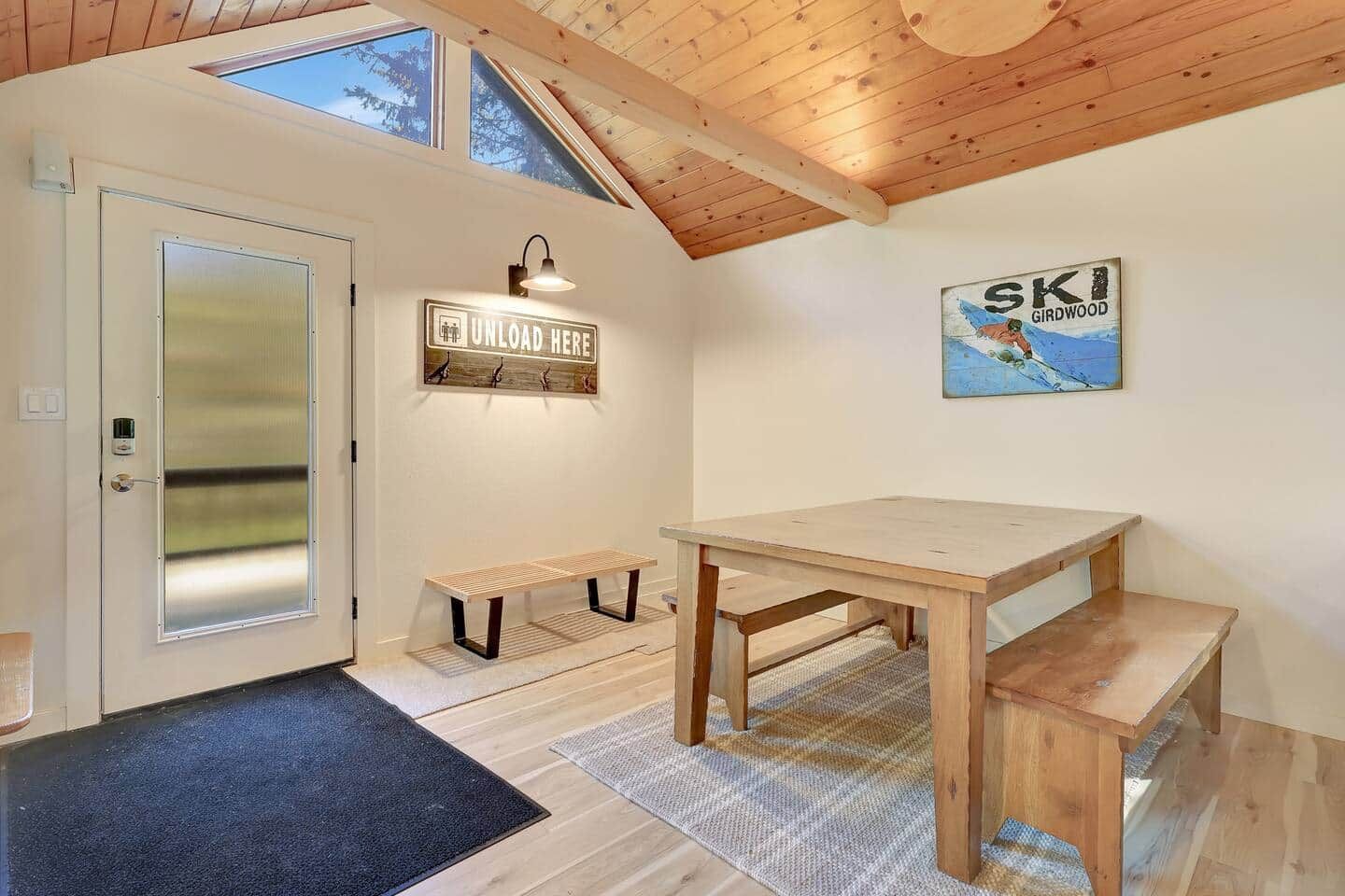 Una habitación con techo de madera, un banco, una mesa, una puerta con ventanal y un letrero en la pared.