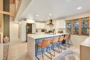 A modern kitchen with a central island, bar stools, and a view of the outdoors through large windows. - Bright & Stylish Girdwood Home- Ski, Bike, Explore (Anchorage)