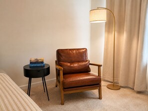 Una habitación con una butaca de cuero marrón, una mesita de noche negra con libros, una lámpara de pie y una cama con cubierta blanca.