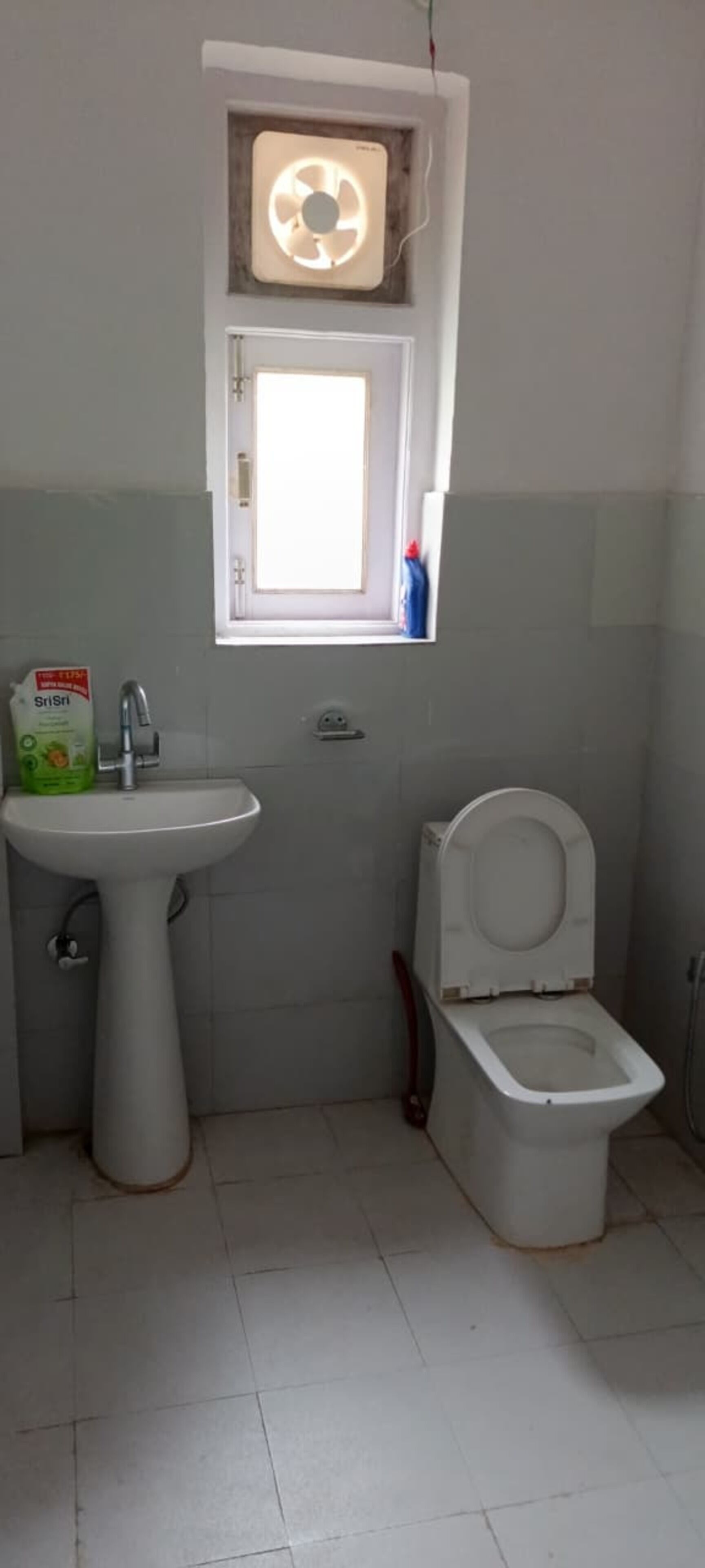 A small bathroom with a pedestal sink, toilet, and a window with a fan.