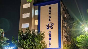 Un hotel llamado 'ROYAL ORBIT' con un letrero vertical llamativo, coches estacionados y un árbol en primer plano.