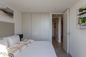 A bedroom with a bed, a bedside table, a lamp, a wardrobe, a door leading to another room, and a wall-mounted shelf with plants. - JEMS PROPERTY (Cape Town)