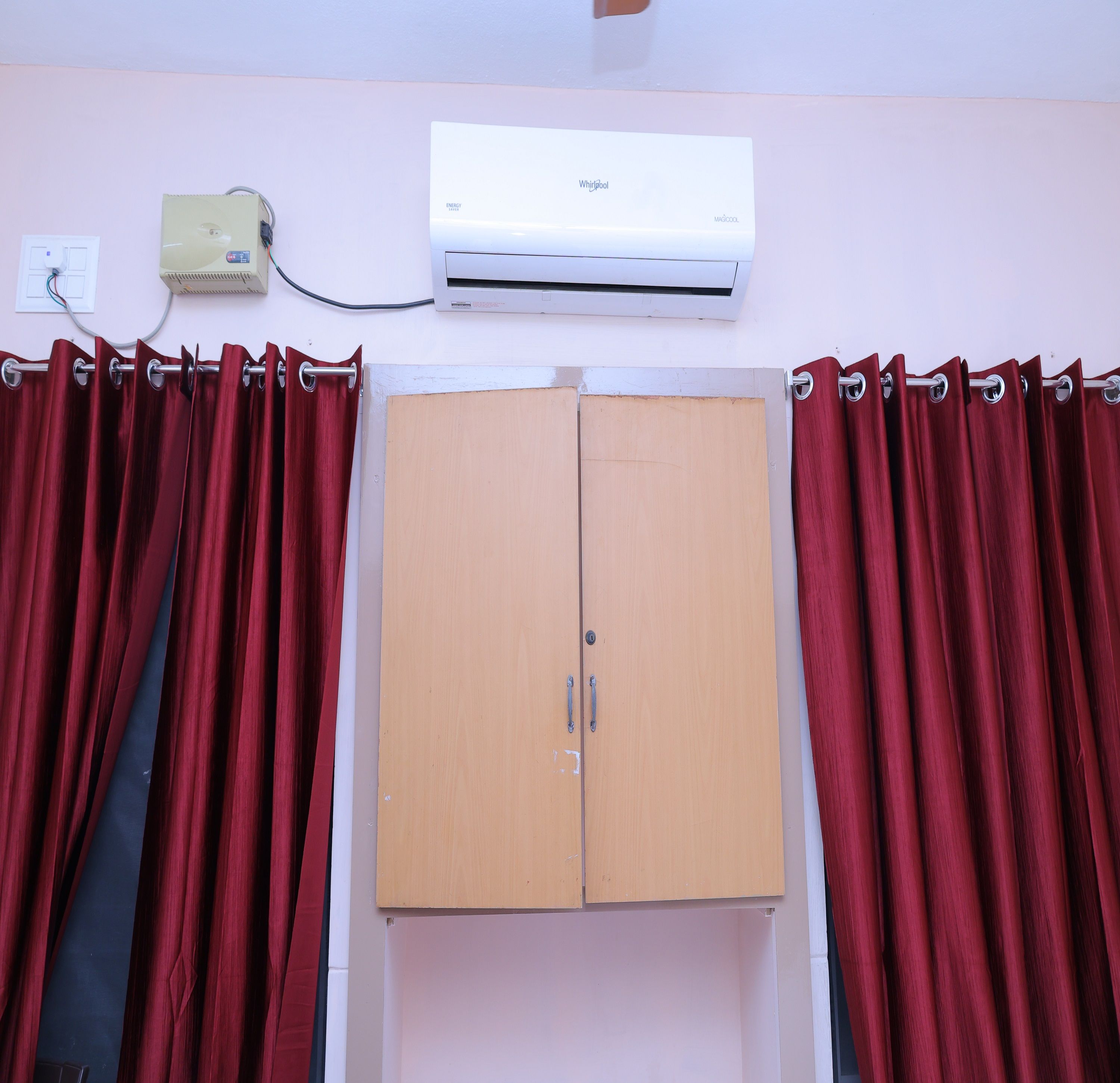 A room with a wardrobe, red curtains, an air conditioning unit, and a wall socket.