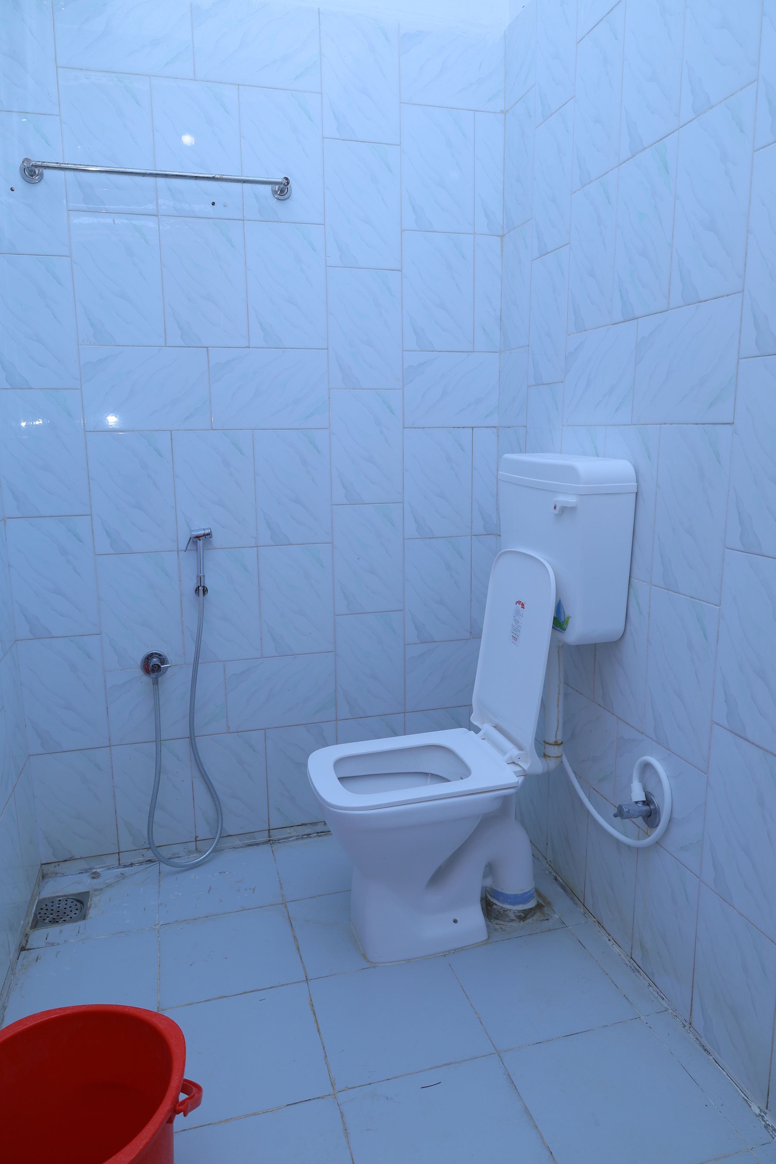 Un baño pequeño con azulejos blancos, que incluye un inodoro, una ducha y un balde rojo en el suelo.