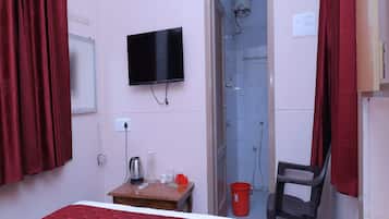 Habitación de hotel con cama, televisor, ducha, mesa con tetera, vasos y un cubo rojo.