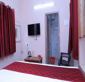 Habitación de hotel con cama, televisor, ducha, mesa con tetera, vasos y un cubo rojo.
