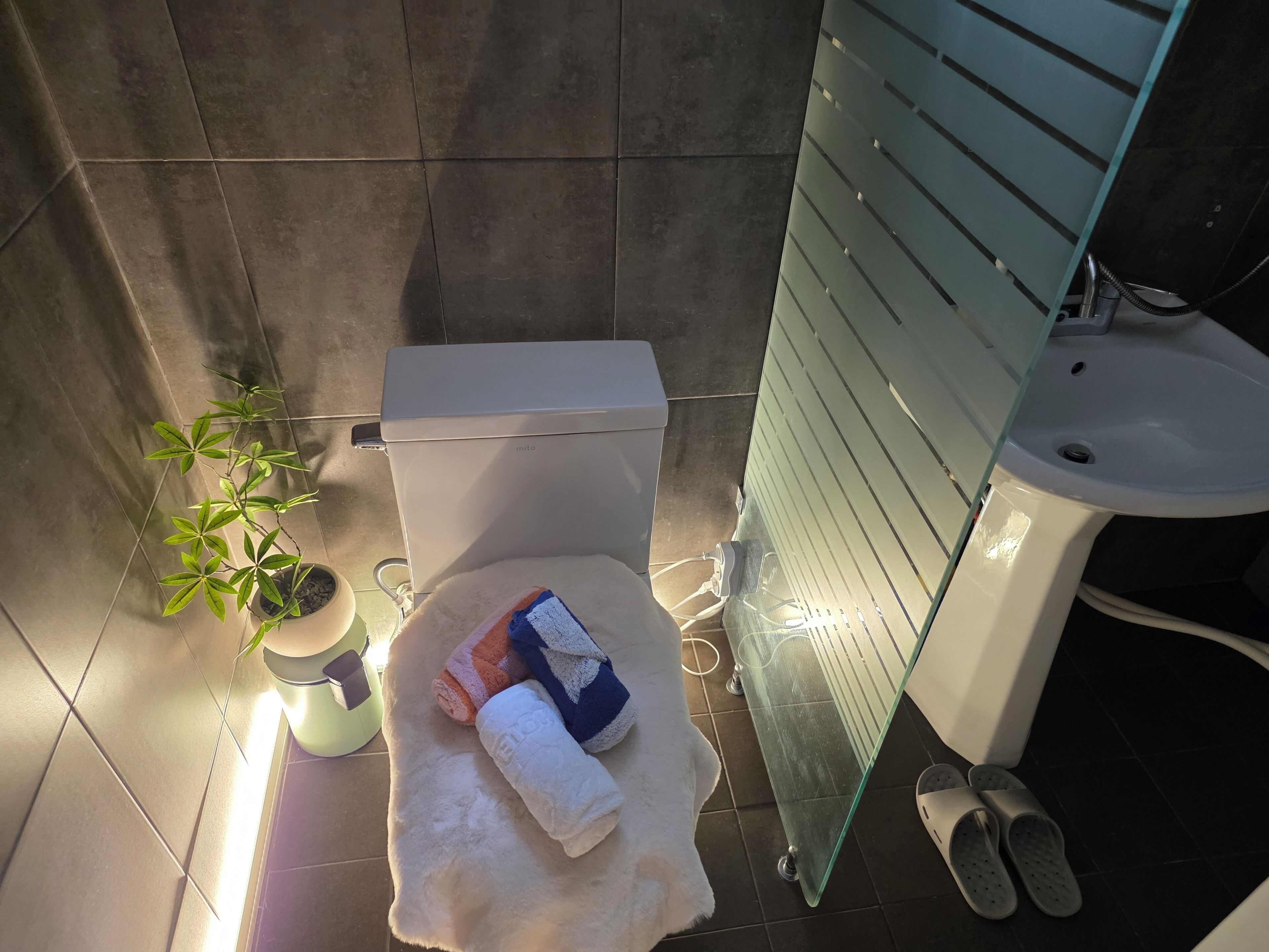 A bathroom with a toilet, a sink, a glass shower enclosure, slippers, and a towel on the floor.