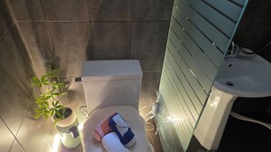 A bathroom with a toilet, a sink, a glass shower enclosure, slippers, and a towel on the floor. - Lumi House Itaewon (Seoul)