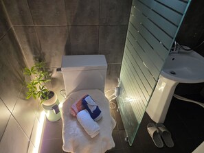 A bathroom with a toilet, a sink, a glass shower enclosure, slippers, and a towel on the floor.