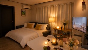 A hotel room with a bed, bedside table with candles, a television, and a dining area with food and drinks. - Lumi House Itaewon (Seoul)