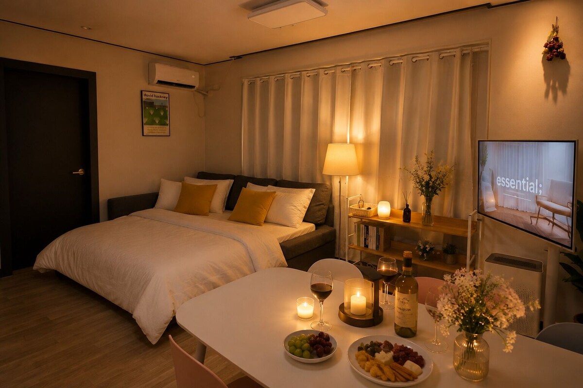 A hotel room with a bed, bedside table with candles, a television, and a dining area with food and drinks.
