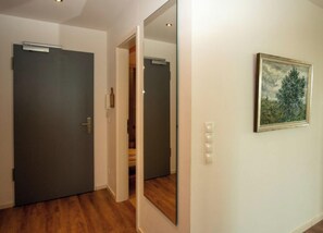 Ein Korridor mit einer dunklen Tür, einem Spiegel der einen Raum reflektiert und einem Bilderrahmen an der Wand.