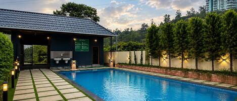 A swimming pool with a covered changing room, a pathway leading to the pool, and a landscaped area with trees.