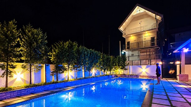 Zona de piscina iluminada de noche con un edificio de dos plantas al fondo.