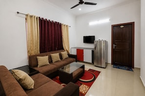 A compact living space with a sofa, a glass coffee table, a flat-screen TV, a refrigerator, and a wooden door. - Casa Valley (Munnar)