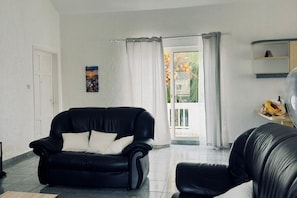 Una sala de estar con un sofá de cuero negro, una puerta blanca, un cuadro en la pared y vistas al exterior a través de una puerta de vidrio.