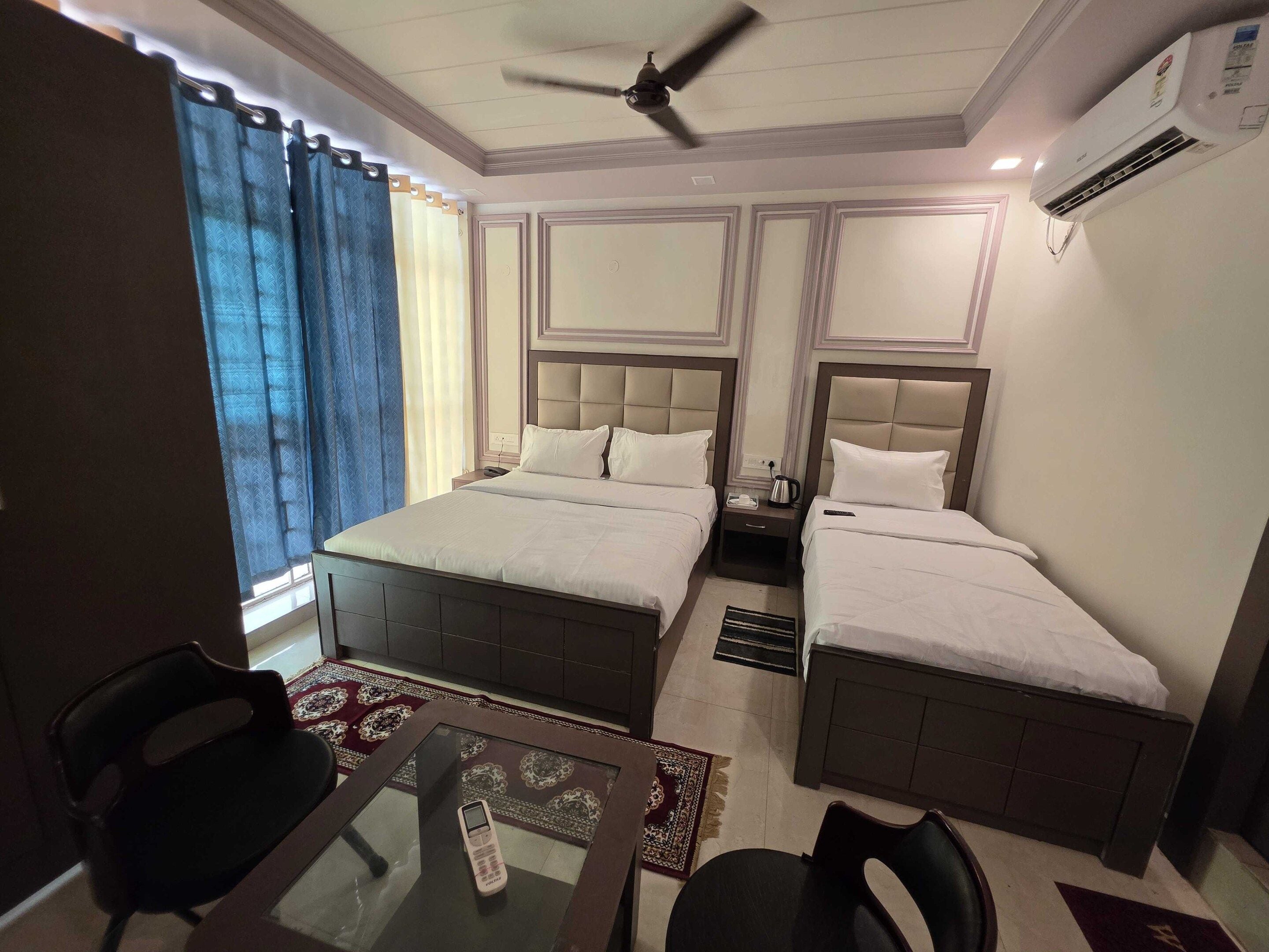A hotel room with a bed, two bedside tables, a ceiling fan, an air conditioning unit, a window with curtains, and a small table with a chair.