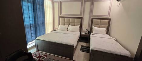 A hotel room with a bed, two bedside tables, a ceiling fan, an air conditioning unit, a window with curtains, and a small table with a chair.