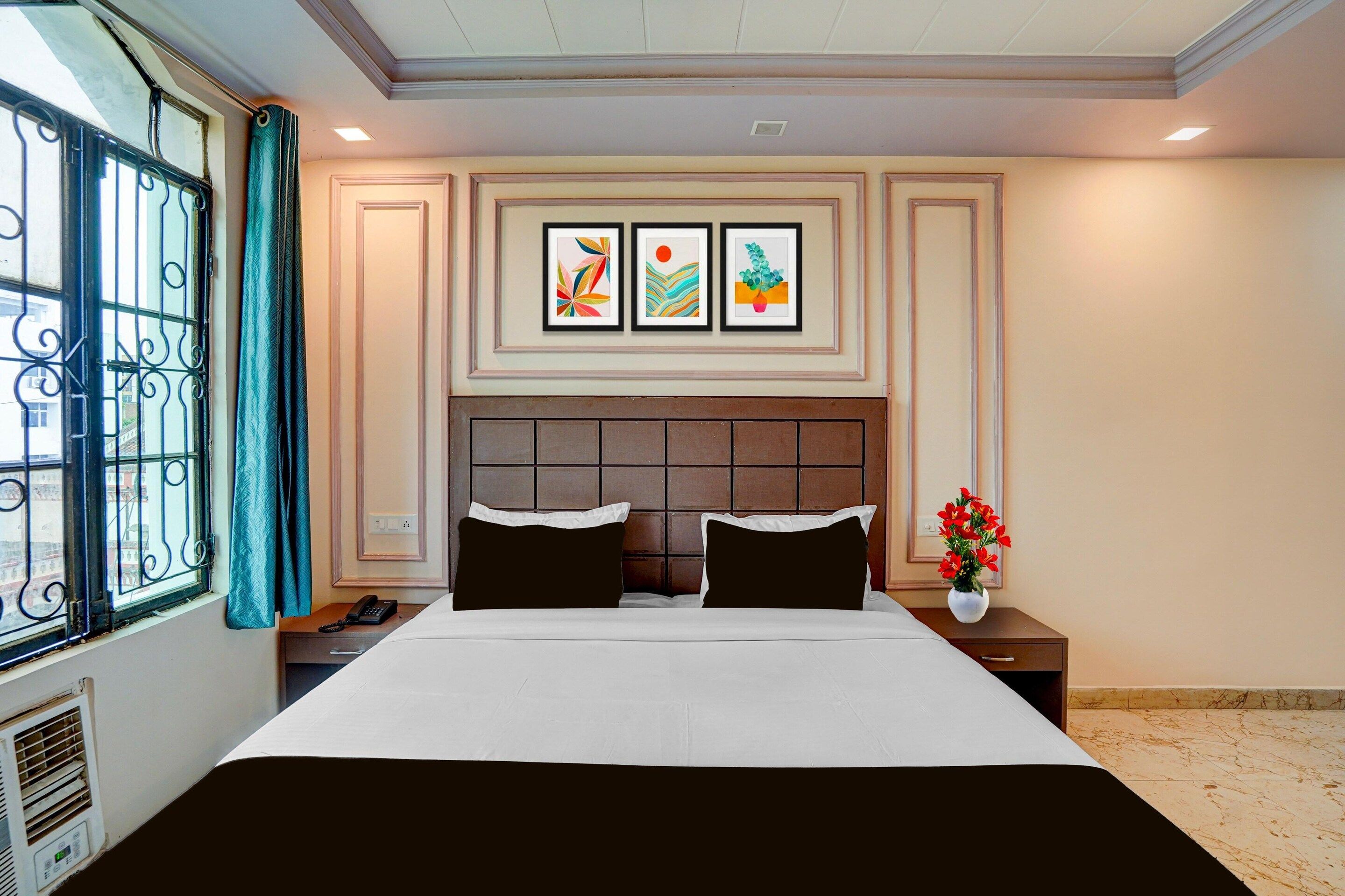 A hotel room with a bed, two pillows, a nightstand, a phone, a vase with flowers, and three framed pictures on the wall.