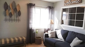 En stue med sofa, bænk, et vindue med gardiner, lampe og et skilt med teksten 'welcome'.