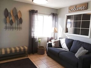 A living room with a sofa, a bench, a window with curtains, a lamp, and a 'welcome' sign. - Great Choice!  One Bedroom Suite!  Close to Shopping, Parks & Museums! (West Des Moines)