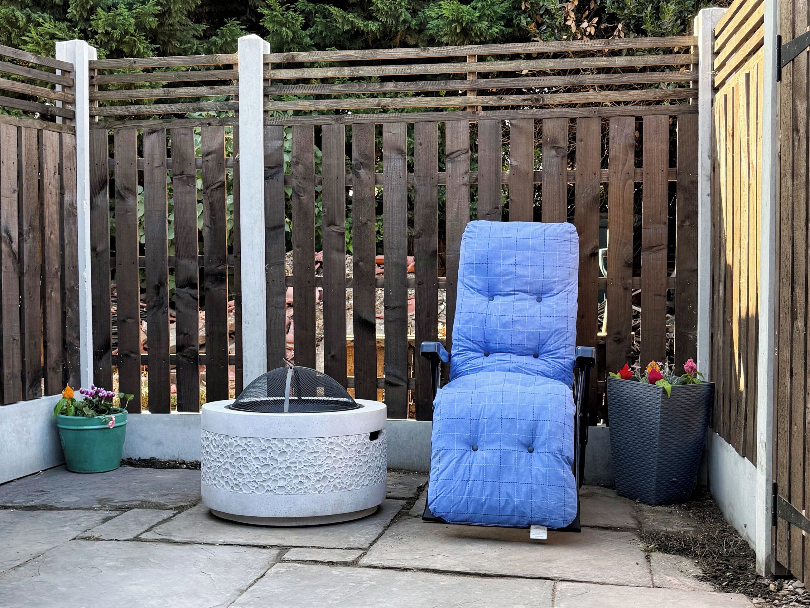 Un espace patio avec un fauteuil bleu rembourré, un chauffage d’extérieur rond et une clôture en bois.