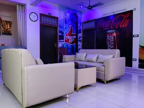 A modern living room with a sofa, armchair, and coffee table. There are posters on the wall, including a Coca-Cola ad. - Shri Shyam Gest House (Sultanpur)