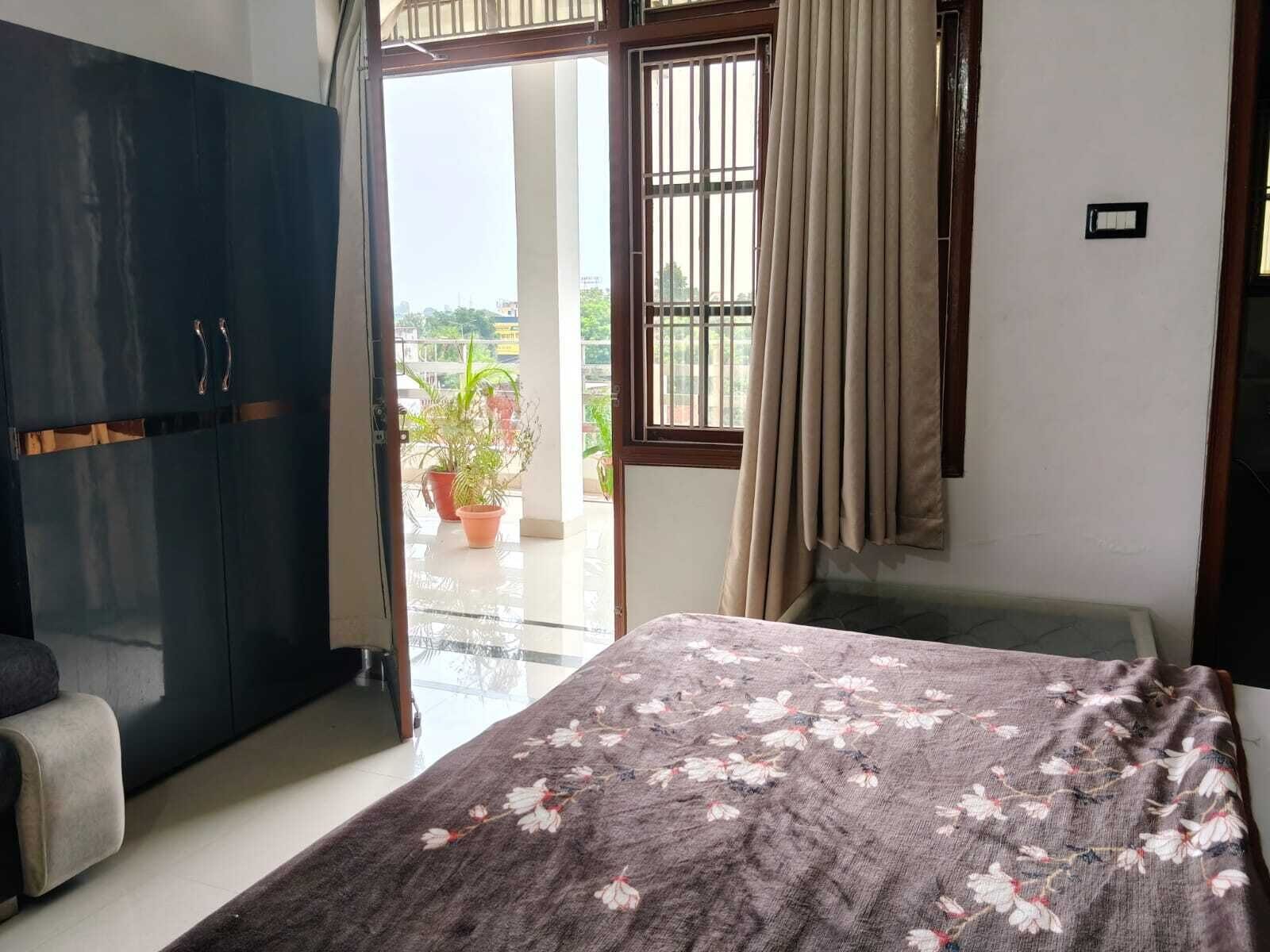 A room with a bed, a wardrobe, a balcony with potted plants, and a view of the outside.