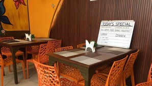 Interior de restaurante con mesas de madera y sillas anaranjadas, un panel con las especialidades del día y un cuadro floral en la pared.