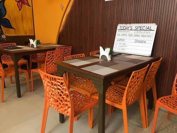 A restaurant interior with wooden tables and orange chairs, a menu board displaying specials, and a wall with a floral painting.