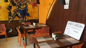 Interior de un restaurante con sillas naranjas, una mesa a cuadros y una pizarra con opciones de desayuno y almuerzo.
