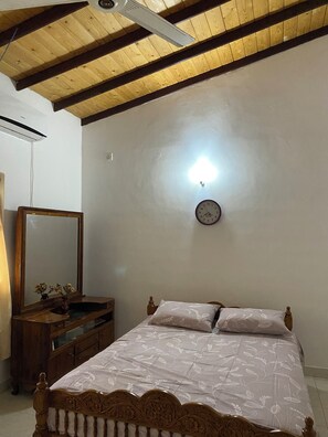 Une chambre avec un plafond en bois, un lit recouvert d’une couvre-lit à motifs floraux, une table de chevet avec un miroir et une horloge murale.