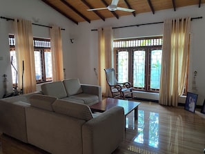 Una sala de estar con un sofá, una mesa de centro de madera y una silla. Hay ventanas con cortinas, un ventilador de techo y vistas a zonas verdes al exterior.