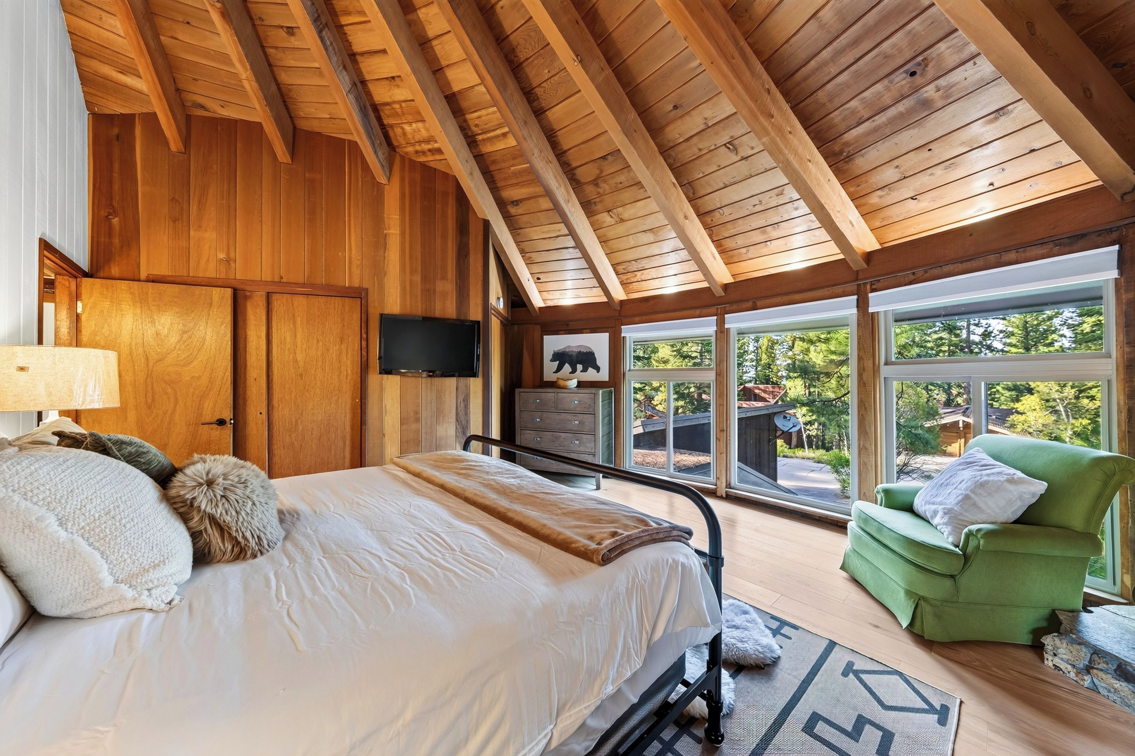 Un dormitorio con techo de madera, una cama con ropa de cama blanca, un sillón verde y vistas al exterior.