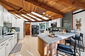 A modern kitchen with wooden beams, a marble island, and a view into the dining area. - Redwood Home | Sauna + Hot Tub | Near Lifts + Village | Roundhouse at Northstar (Truckee)