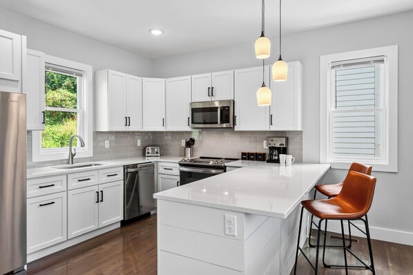 A modern kitchen with white cabinets, a stainless steel refrigerator, a microwave, and a large island with bar stools. - Blue Ridge Views + Balcony + Two Living Rooms | Downtown | Tranquil River Home (Asheville)