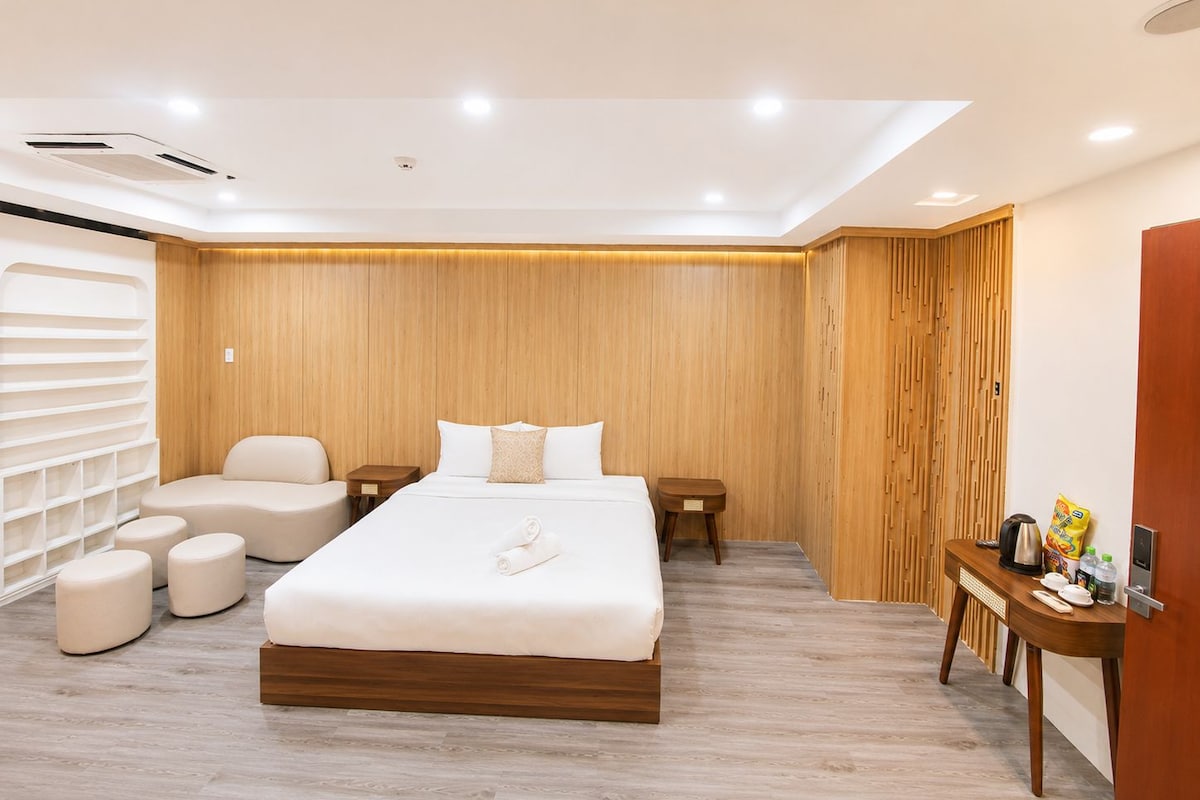 A modern bedroom with a large bed, two bedside tables, a wooden wardrobe, and a seating area with ottomans.