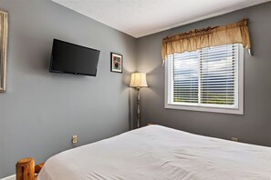 Ein Schlafzimmer mit einem Bett, einem flachbildfernseher an der Wand, einem Fenster mit Jalousien und einer Lampe.
