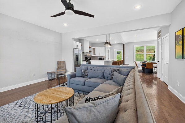 A modern living room with a sectional sofa, a coffee table, and a dining area with a table and chairs. - French Broad River Views • Balcony • Near Silver Line Park | Riverside House (Asheville)