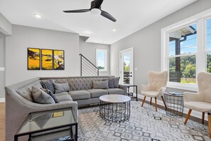 A modern living room with a grey sofa, two armchairs, a coffee table, and a ceiling fan. - French Broad River Views • Balcony • Near Silver Line Park | Riverside House (Asheville)