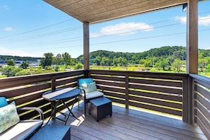 A wooden deck with outdoor furniture, overlooking a green landscape and a body of water. - French Broad River Views • Balcony • Near Silver Line Park | Riverside House (Asheville)