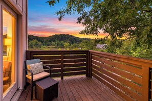 A wooden deck with a wicker chair and a cushion, overlooking a forested landscape with a sunset. - Balcony + River Views + Full kitchen | River Arts District | Sage House (Asheville)